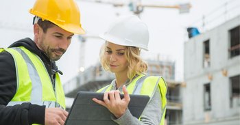 Eine Frau und ein Mann mit Bauhelmen stehen auf einer Baustelle und schauen gemeinsam auf ein Tablet