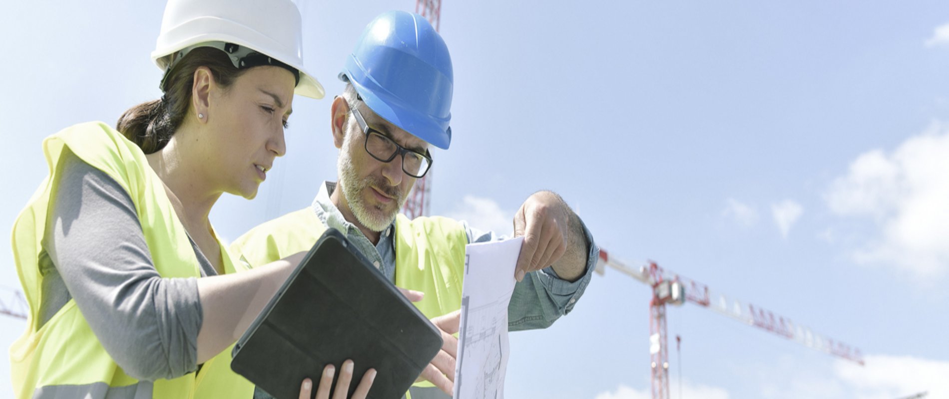 Eine Frau und ein Mann mit Bauhelmen stehen auf einer Baustelle und schauen prüfend auf ein Tablet und einen Bauplan