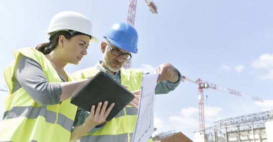 Eine Frau und ein Mann mit Bauhelmen stehen auf einer Baustelle und schauen prüfend auf ein Tablet und einen Bauplan