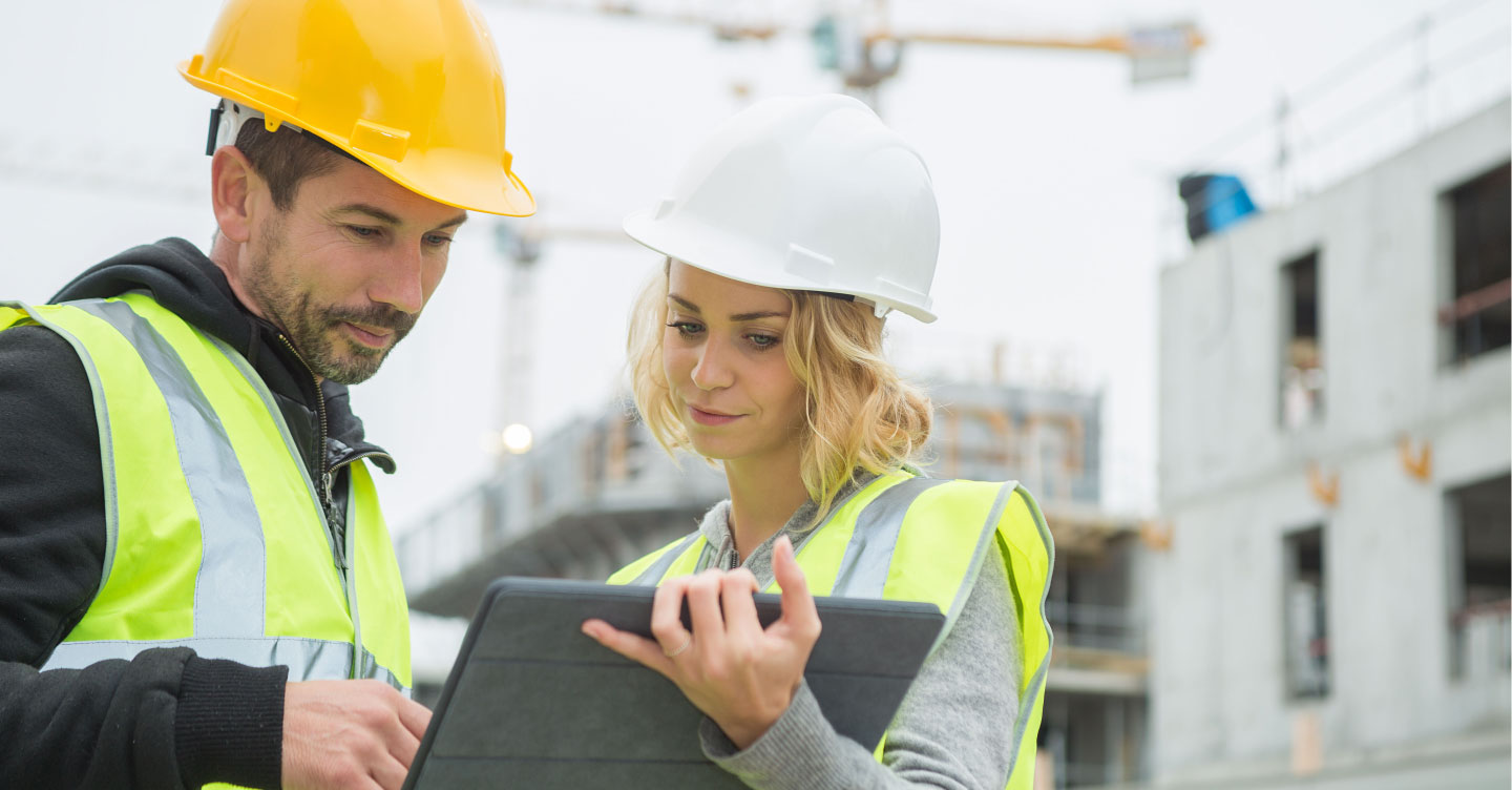 Eine Frau und ein Mann mit Bauhelmen stehen auf einer Baustelle und schauen gemeinsam auf ein Tablet